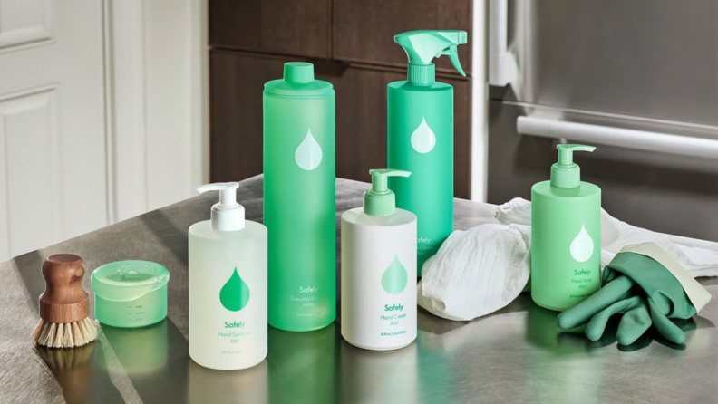 Product shot of green and white Safely product line on counter next to scrubbing brush and green rubber gloves.