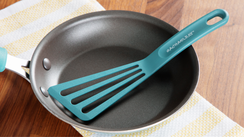 Agave blue colored plastic spatula sitting inside of small nonstick frying pan on top of dish cloth and wooden countertop surface.