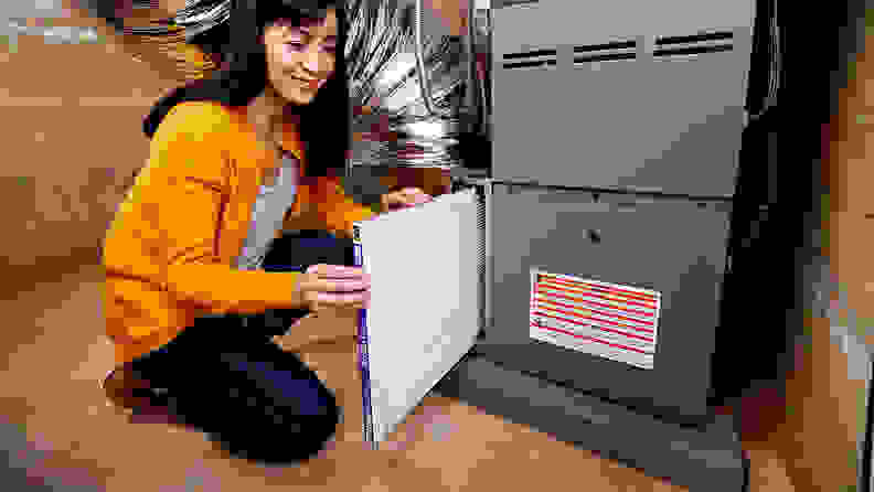 A woman kneels beside a furnace, changing in a new Filtrete Micro Allergen Defense Air Filter.