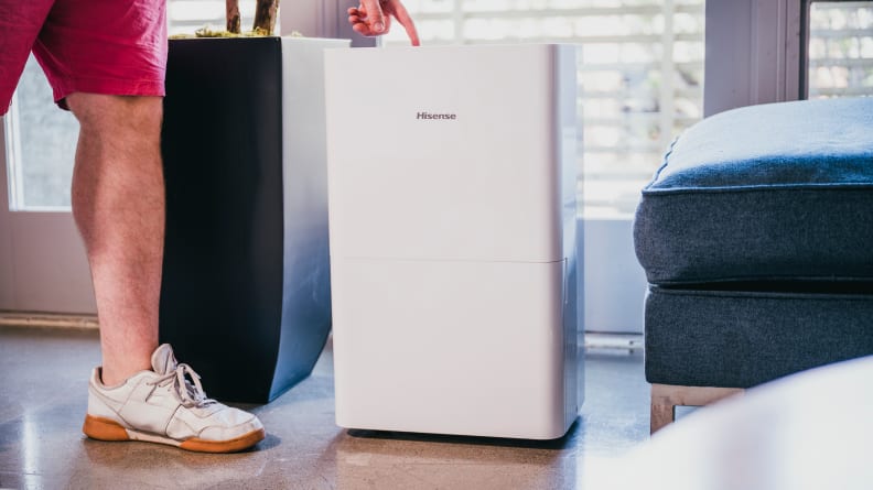 A person wearing red shorts steps to a white Hisense DH7021K1W dehumidifier and pushes settings on the top.
