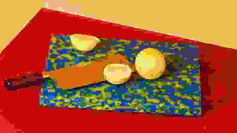 A multi-colored plastic cutting board sits on a red surface with halved lemons and a large cleaver perched on top.