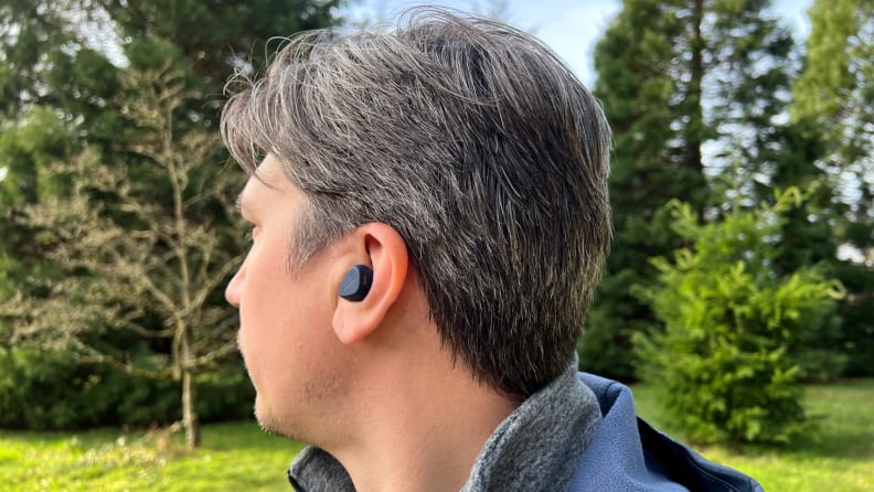 An earphone sits in the ear of a brown and white haired man in a park with trees and grass.
