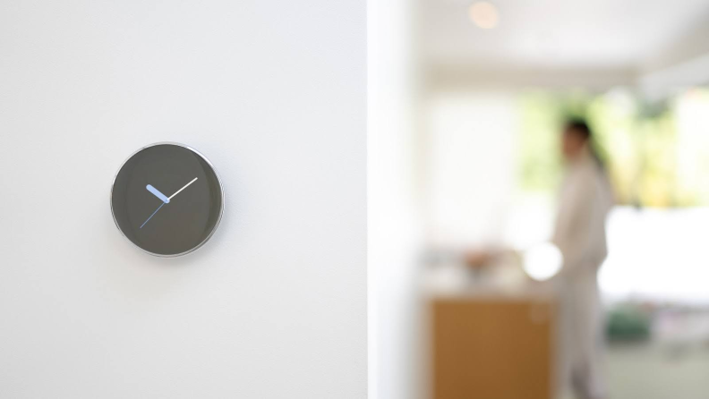 The fourth-gen Nest Learning Thermostat hangs inside of a home on a white wall with the clock face displayed on the digital screen.