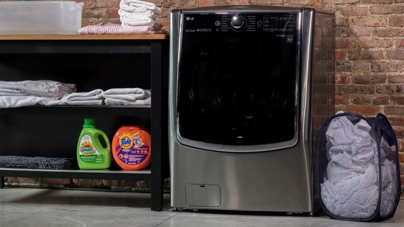 A grey LG WM9000HVA washer sits in front of a brick wall next to a clothes hamper.