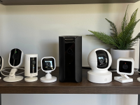 Eight smart security cameras from various brands sitting on a wood shelf with a faux plant in the background
