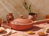 The terracotta-colored Always Pan is in the center of the image. The color is earthy with an orange hue. Next to the pan, pottery vases, drinkware, and plates of nuts and snacks are on display..