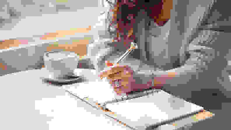 Young woman in blue warm sweater sitting near the big window of coffee shop and writing a Christmas shopping list with cup of blue latte. Planning Christmas holidays. Organising and planning concept.