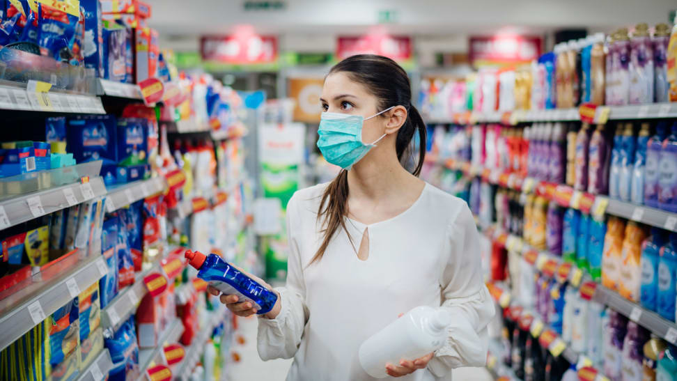 A woman, wearing a face mask, is in the cleaning product aisle of a store, comparing two different options.
