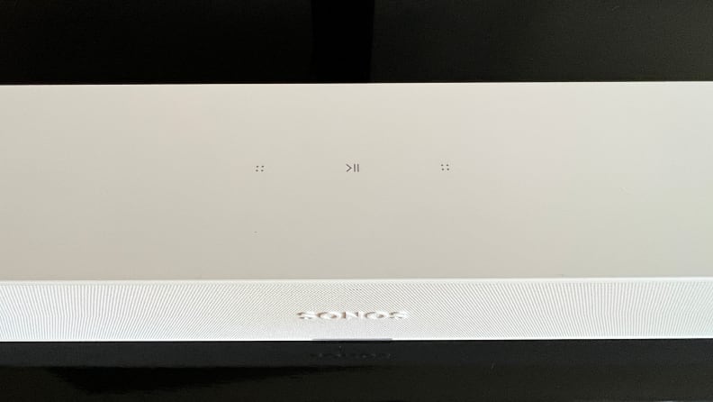 The white Ray soundbar's top touch keys are shown as it sits on a black and brown console.