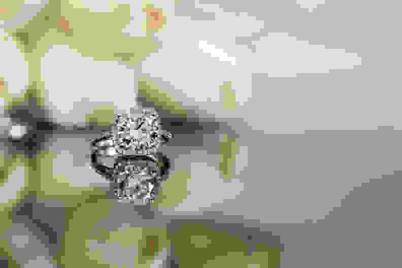 An engagement ring with a halo setting with white flowers behind it, something to consider before buying an engagement ring.