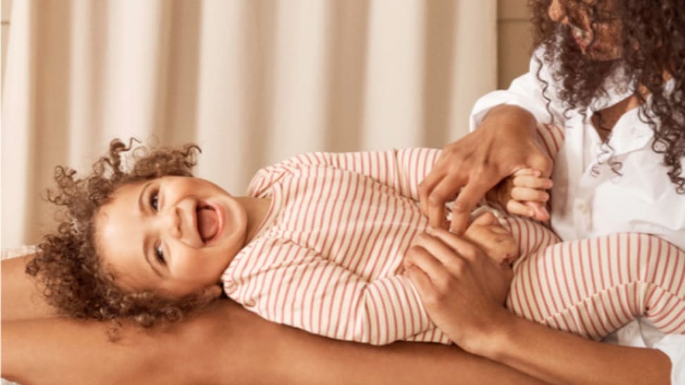 A baby laughing as they are being tickled by their mom, while wearing a Maisonette red striped one piece.