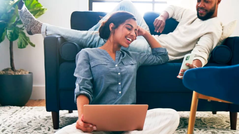 An image of a woman seated on the floor, holding a laptop and showing a man seated above her on a sofa, as he leans down to show her something on his phone.