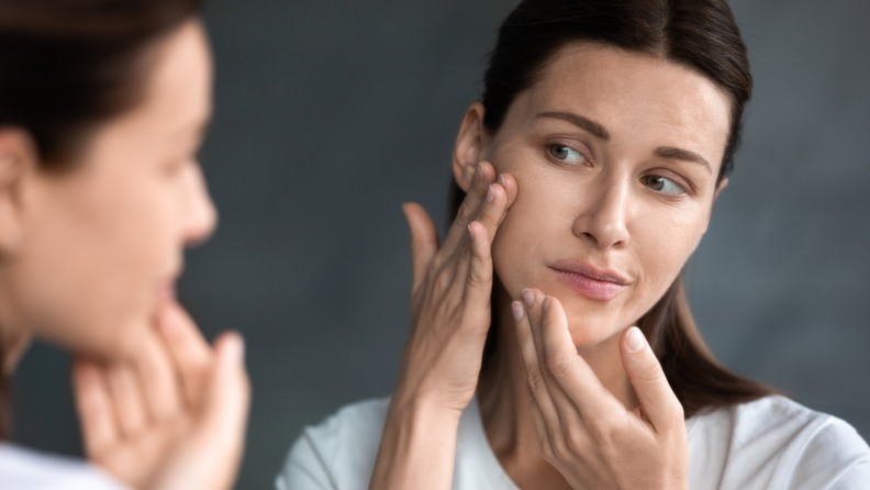 A woman in the mirror with her hands on her cheek checking for acne and other skincare concerns