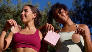 Two women wearing activewear hold a box and a pair of Cakes nipple covers outdoors in the sunlight.