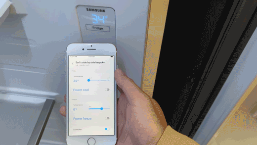Person holding smartphone uses the Samsung SmartThings app to adjust the temperature settings on the Samsung RS28CB7600 side-by-side Bespoke refrigerator.