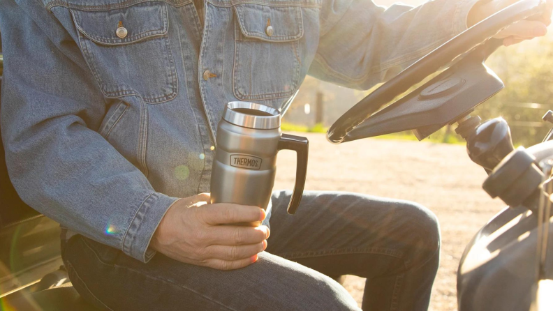 A man driving a tractor holding a Thermos coffee mug.