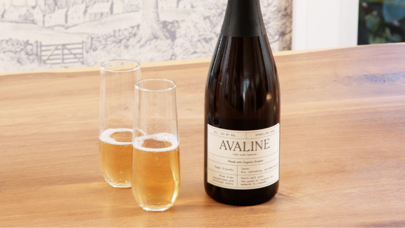 A bottle of Avaline sparkling wine with two glasses filled next to it sit on a wood table.