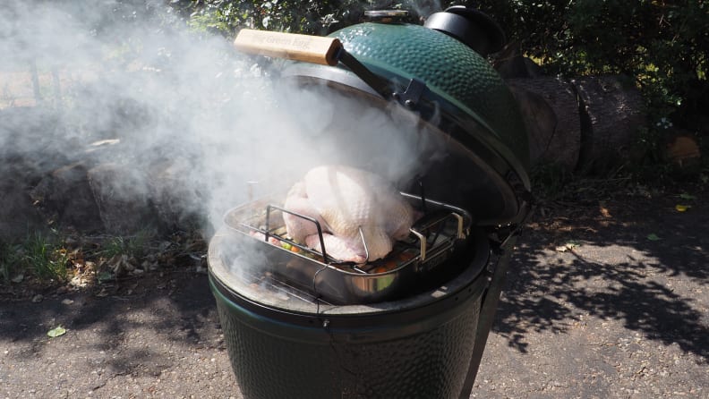 A turkey in a roasting pan is perched on the grill of a Big Green Egg smoker.