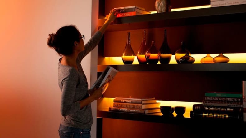 A woman reaches up to adjust the top shelf of a shelving unit with the Philips Hue Bluetooth Lightstrip Plus illuminated in red and yellow in the background