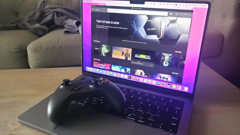 Shot of the Macbook Pro M1 with a gaming controller siting on the keyboard while the monitor displays a collection of digital video game titles.