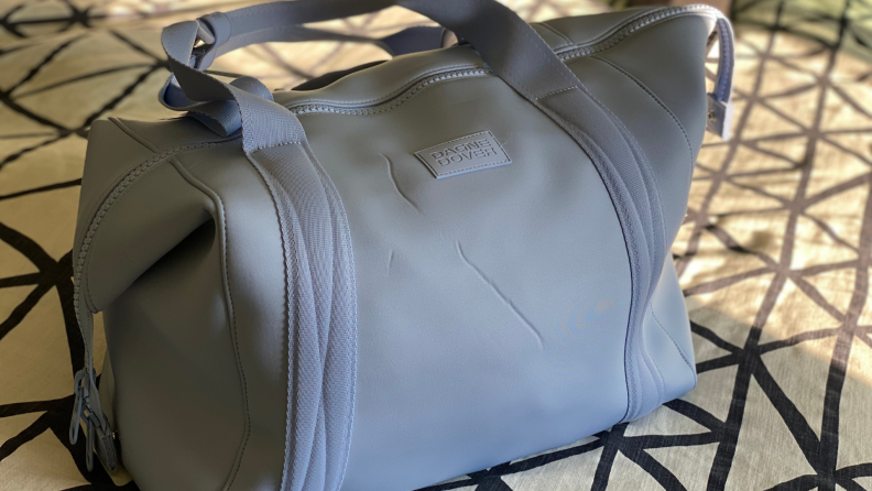 A lavender Landon Carryall on a bed with a geometric bedspread. The photo highlights creasing in the neoprene exterior.