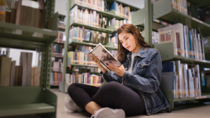 Student sitting on floor of the library, leaning against bookshelf and reading  a textbook