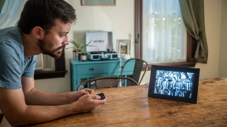 A person leans over a table to point a remote at a digital picture frame.