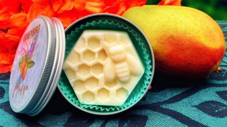 Product shot of cream colored, honey comb shape lotion bar in green tin, next to orange mango and orange flower.