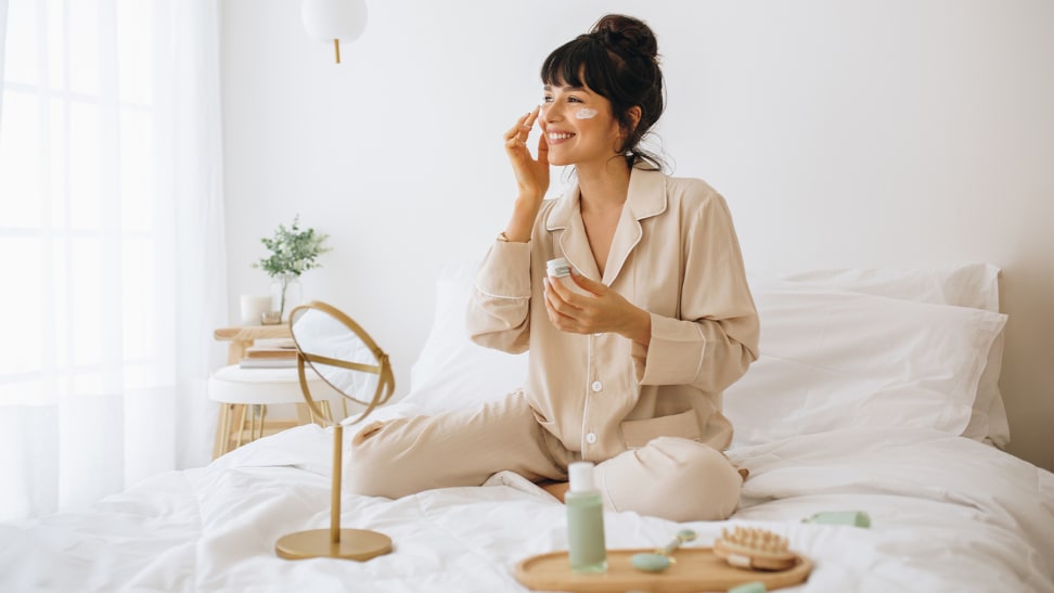 A person sitting on their bed with a tray of skincare and a mirror in front of them.