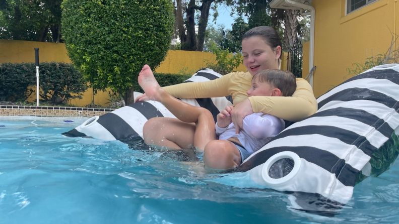 Two children embrace each other on top of black and white pool lounger in water.