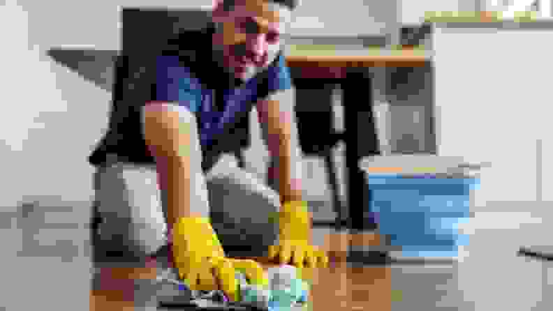 Person down on knees while using yellow rubber gloves and a bucket to clean floor.