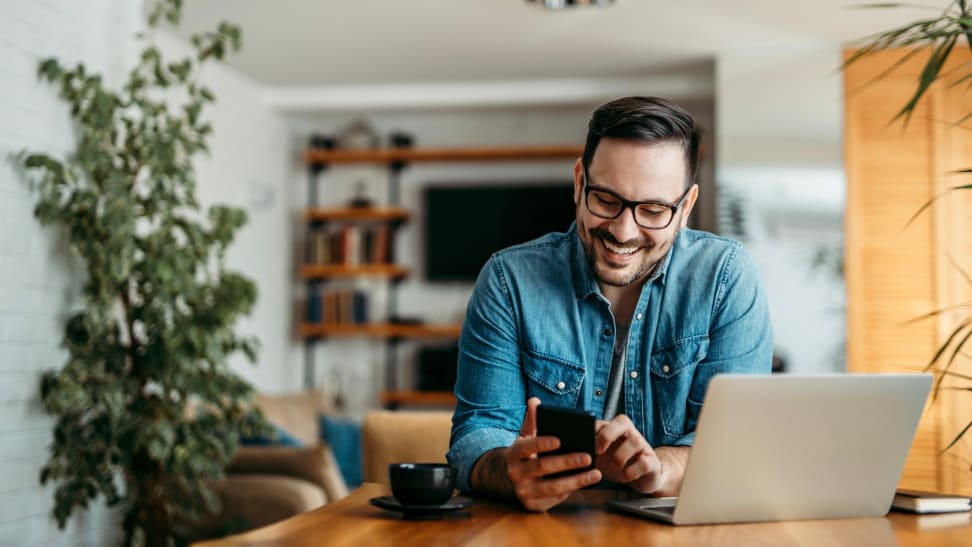 A man smiling while looking down at his phone, while seated in front of his laptop.