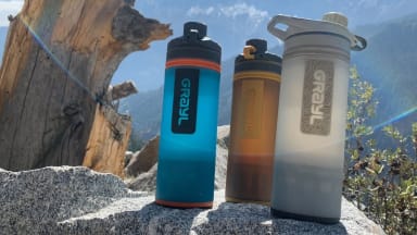 Three Grayl Geopress water bottles sit on a rock with a view of the Indian Himalayas in the background, a log rises on one side, while a lens flare crosses the image.