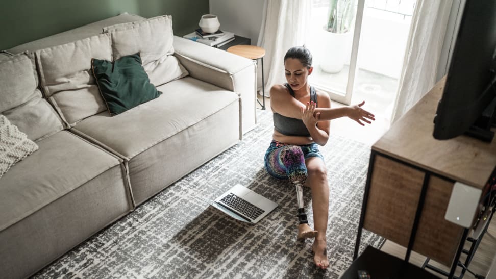 Person with prosthetic leg stretching on living room floor in front of laptop screen.