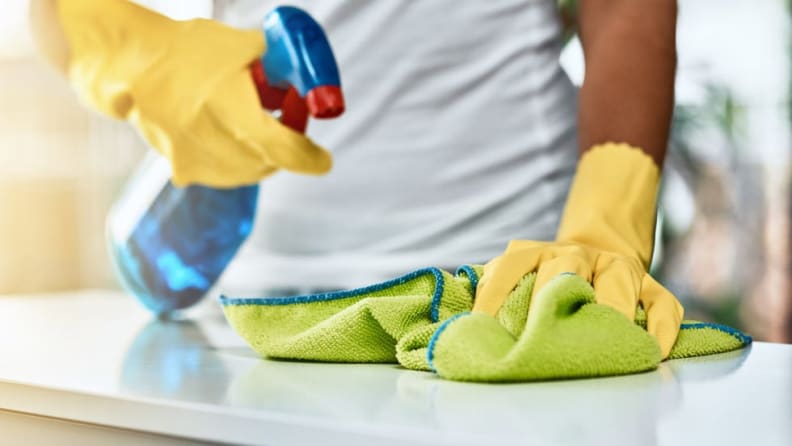Person wearing yellow rubber gloves to wipe countertop using cleaning solution and microfiber cloth.