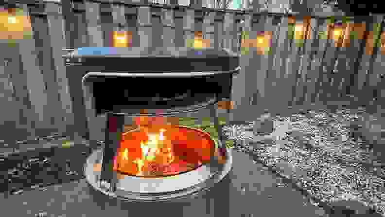 Solo Stove Pi Fire pizza oven with open flame outdoors in front of wooden fence with string lights.