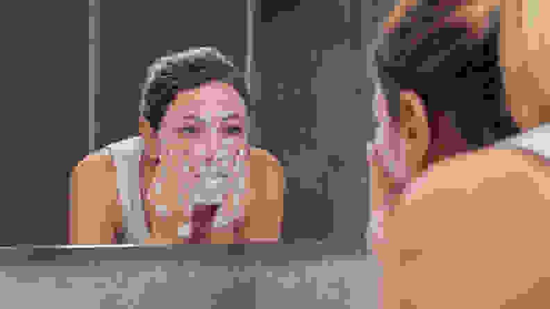 A woman washing her face over the sink and looking in the mirror with foam on her face.
