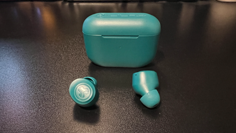 Aqua green earbuds sit in front of a small charging case on a black matte desk.