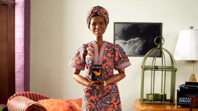 Maya Angelou inspired Barbie dressed in orange and navy kente cloth outfit with matching head wrap while holding book and smiling.