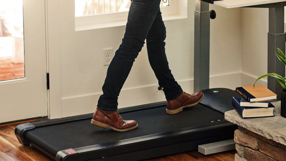 Close-up of a black under-desk treadmill in use at a standing desk in the corner of the room. There is a stack of hardcover books beside it.