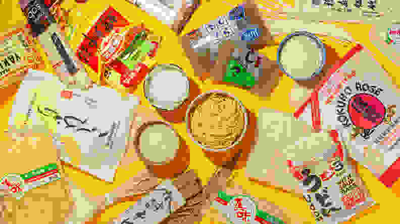 On a yellow table, there are various types of instant ramen, luosifen, udon noodles, white rice, and more scattered around.