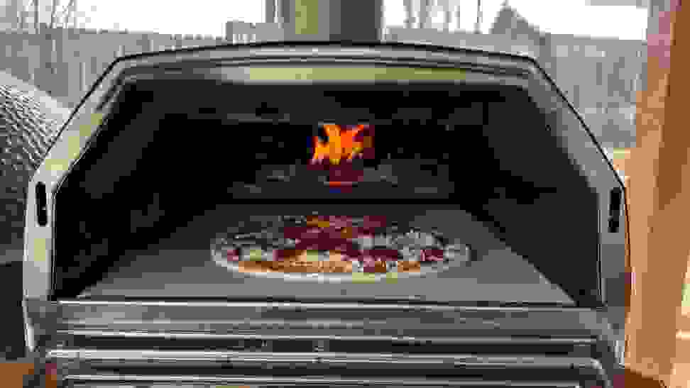 A pizza ready to be cooked sits inside a fired up outdoor pizza oven.