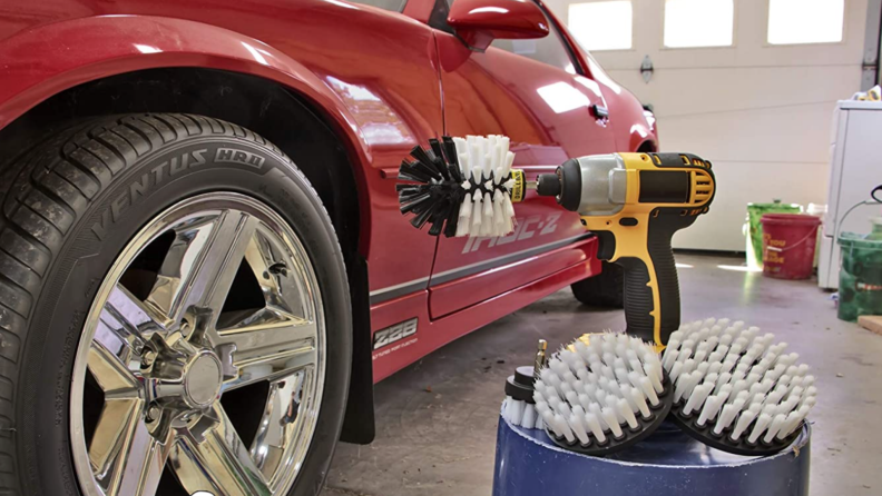 Cordless drill sitting on top of bucket next to brush attachment pieces adjacent to red sports car.