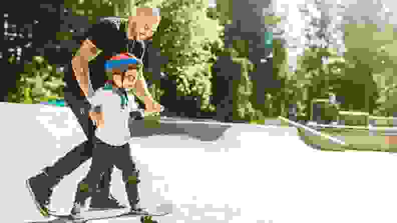 A father smiles as he teaches his young boy to skateboard, holding his arms for balance.