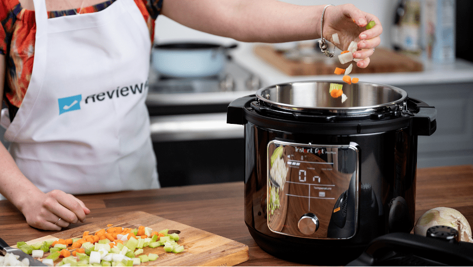 A woman wearing a Reviewed apron putting some chopped carrots, celery, and onion into an Instant Pot pro. There is a stove with a blue pan behind her and some more chopped vegetables on a cutting board in front of her.