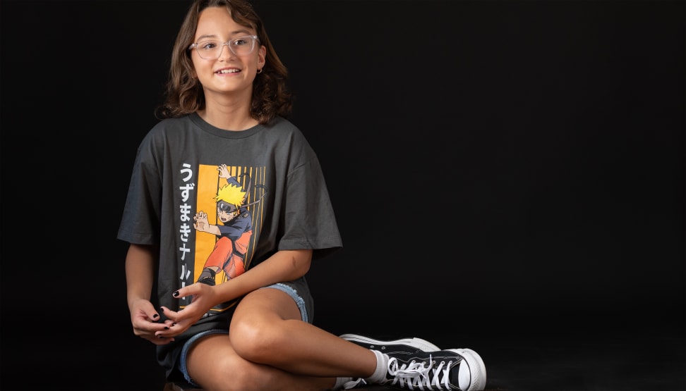 A tween, wearing a Naruto T-shirt and blue denim shorts from Stitch Fix's The Commons, sits on the floor with a black background.