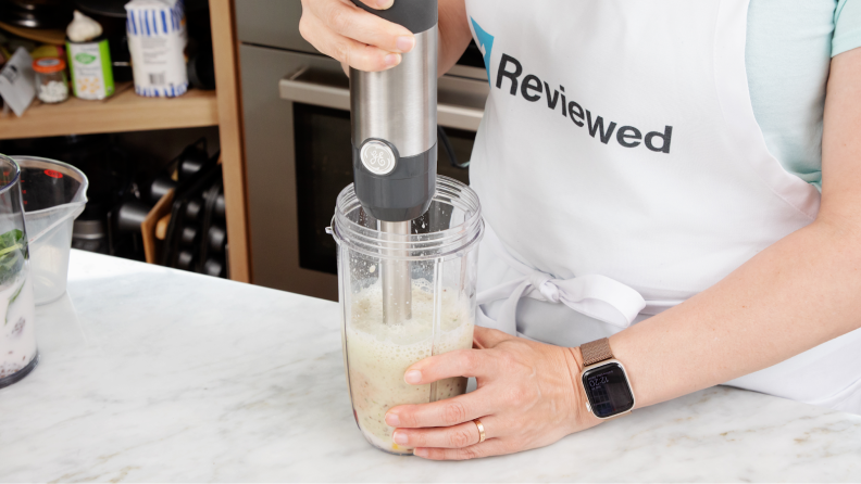 人与围裙站在厨房countertop while using GE Immersion Blender to blend smoothie ingredients inside of plastic cup.