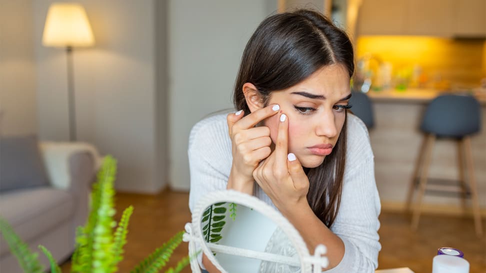 A person looking into a small circular mirror and squeezing a pimple on their cheek.