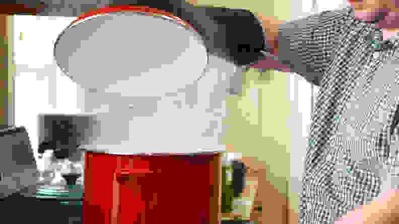 A person is lifting up the lid of a large red stock pot with his mittens on.