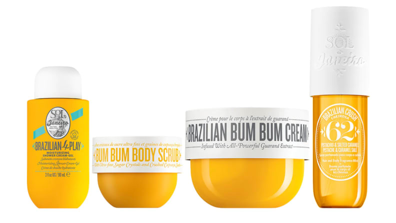 A bottle of body wash, a tub of body scrub, a tub of moisturizer, and a body spray sit side by side in yellow containers.